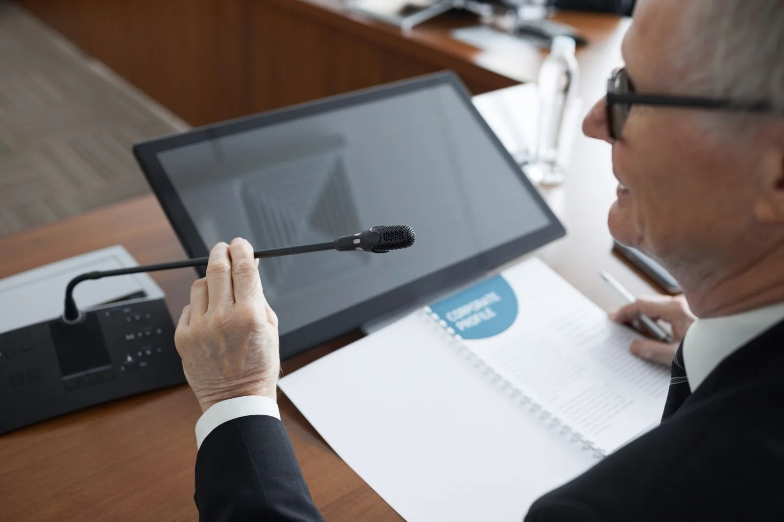Executivo fala ao microfone numa mesa de conferência, com documentos à frente